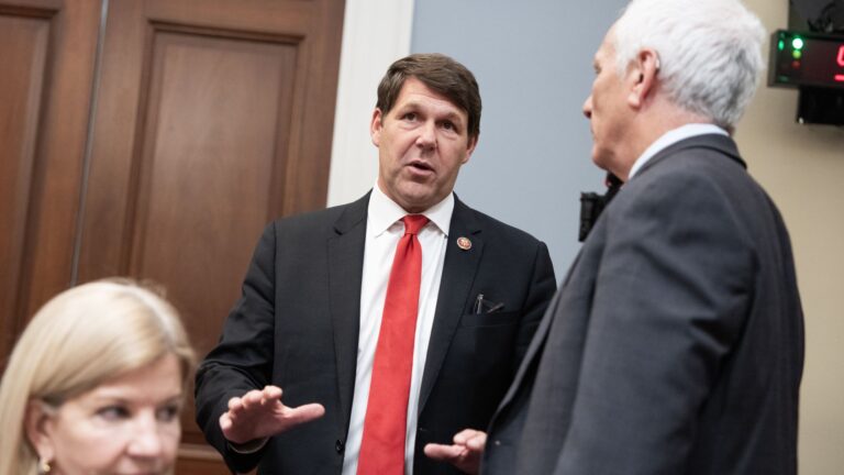A split-screen graphic or photograph: on one side, a military aircraft or drone representing the conflict in Iran; on the other, a medical stethoscope or a hospital sign. The center features a "downward" red arrow over the healthcare side and an "upward" green arrow over the military side to symbolize the shifting budget priorities.